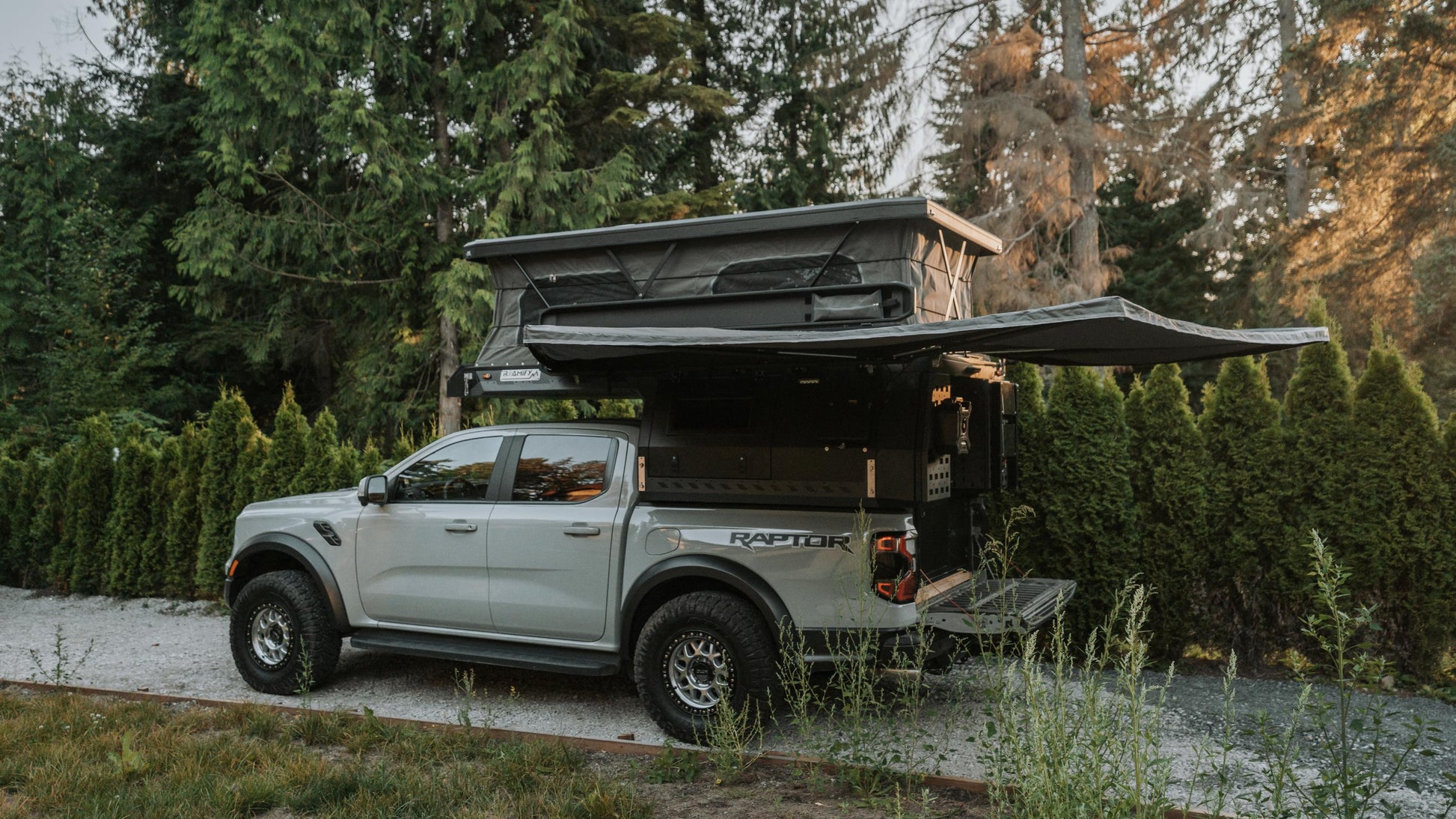White Ford Raptor truck with an open rooftop tent parked in a forested area.