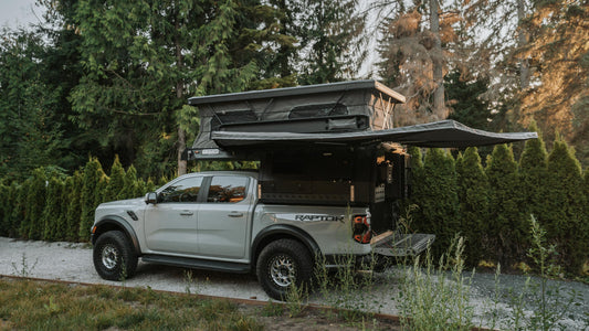 White Ford Raptor truck with an open rooftop tent parked in a forested area.