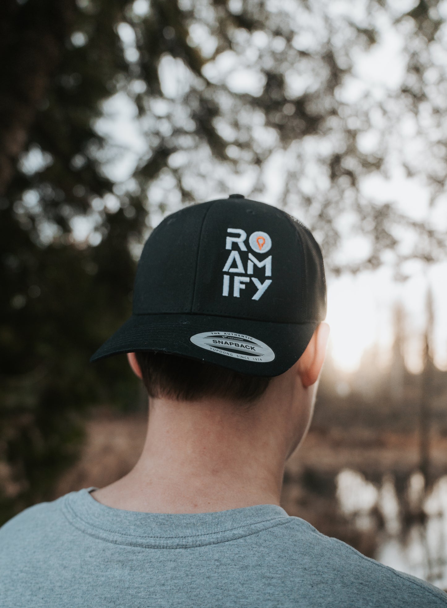 Person wearing a black cap with 'ROAMIFY' text, standing outdoors with trees and water in the background.