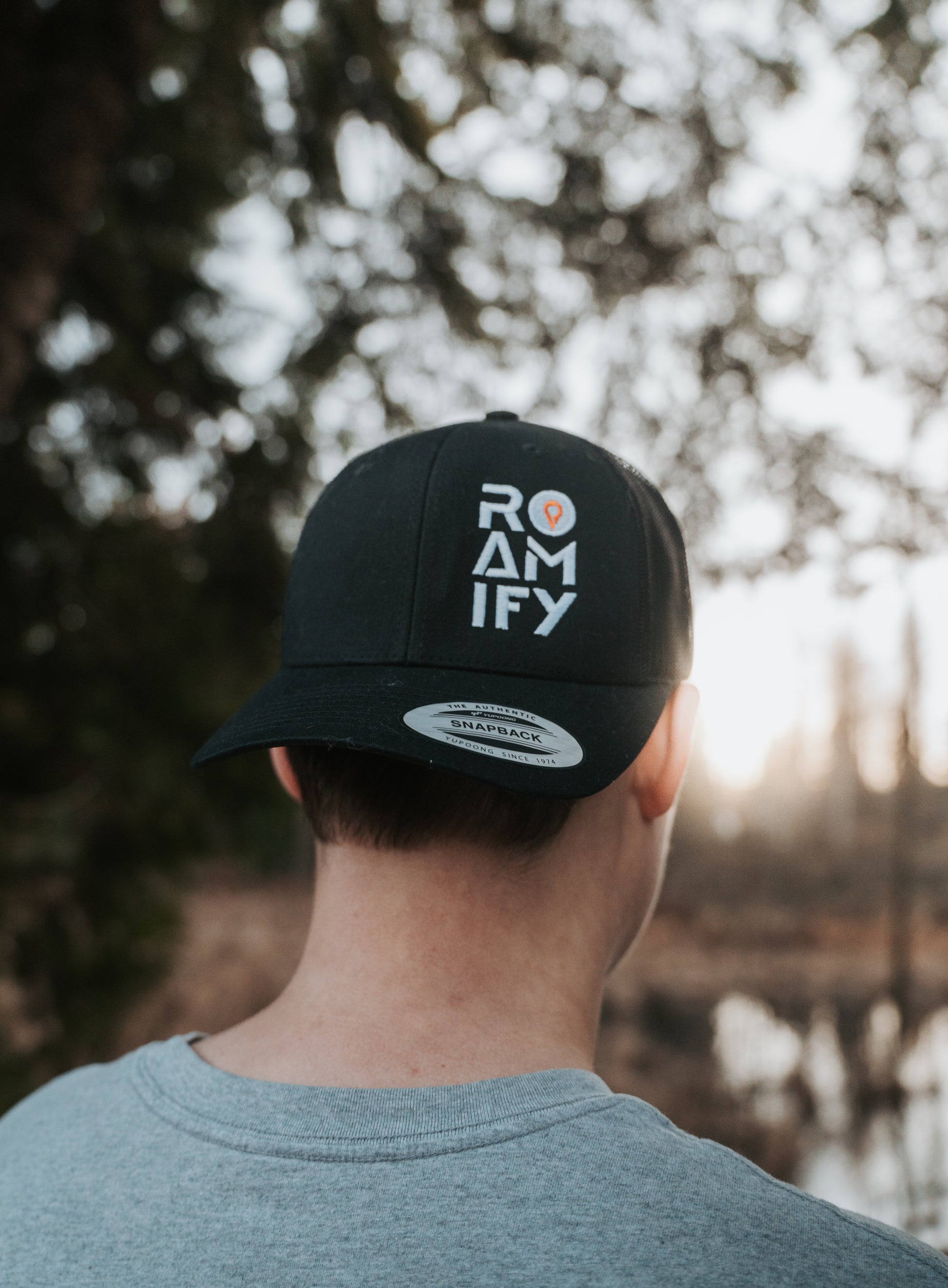Person wearing a black cap with 'ROAMIFY' text, standing outdoors with trees and water in the background.