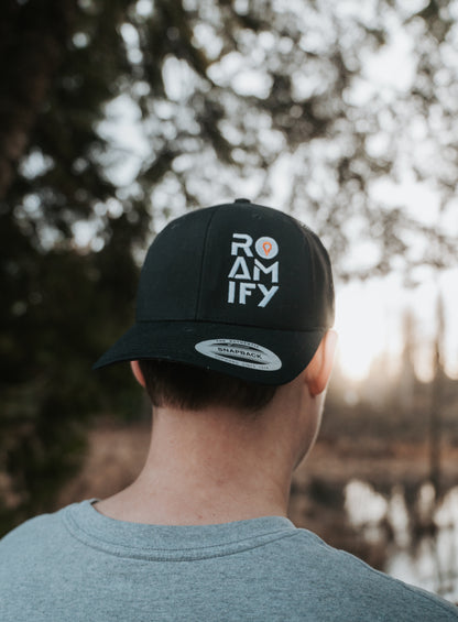 Person wearing a black cap with 'ROAMIFY' text, standing outdoors with trees and water in the background.